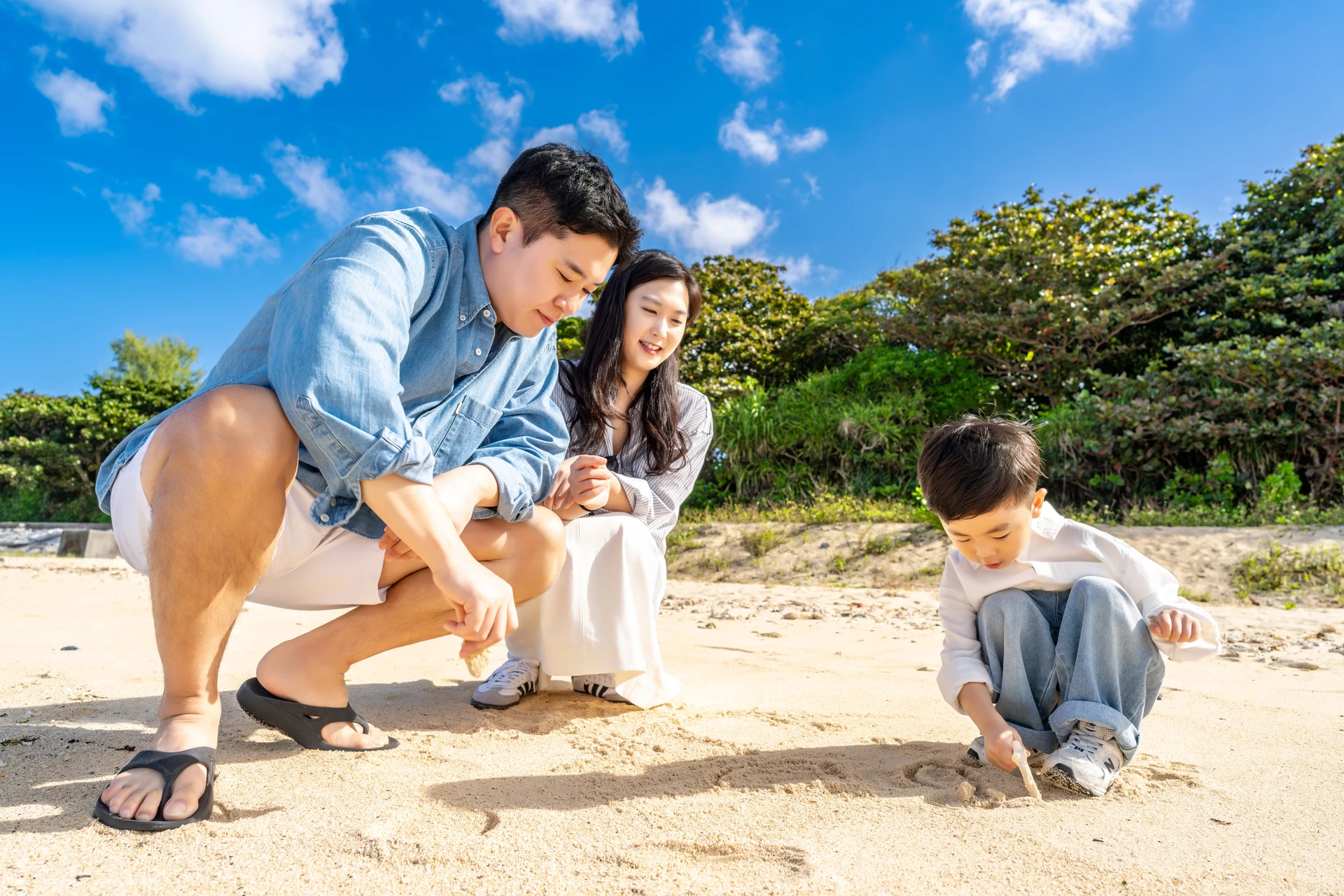 모래사장에 글씨를 쓰는 아이 옆에 쪼그려 앉은 아빠와 엄마