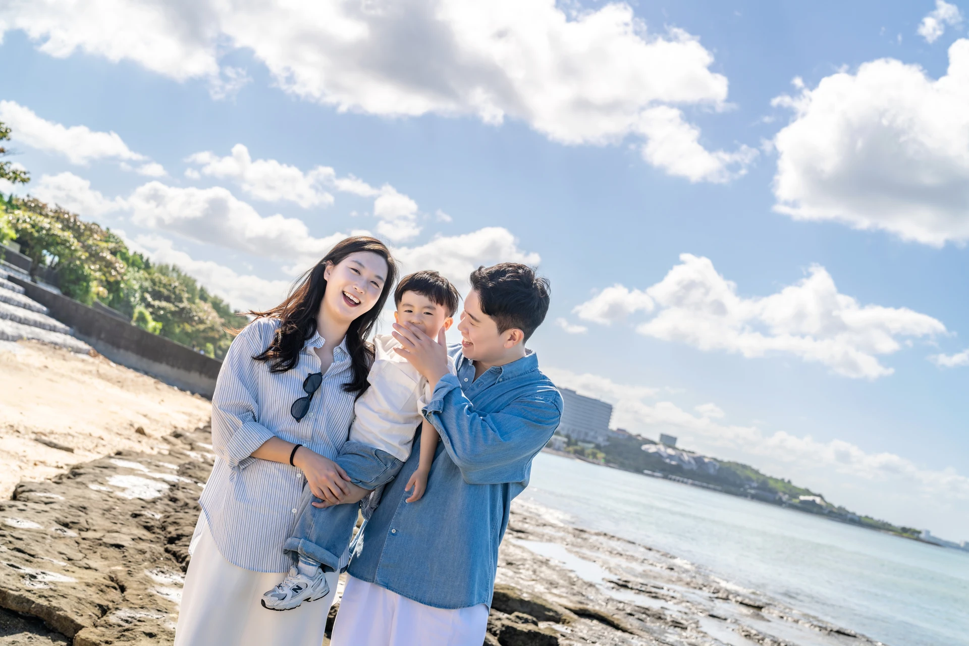아이를 안고 웃는 엄마와 아빠의 클로즈업, 뒤로 보이는 바다와 해변